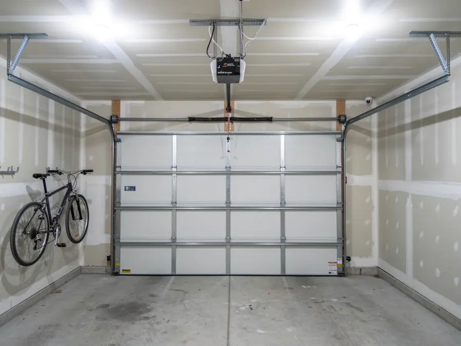 A white garage door is closed in a garage with a bicycle hanging on the wall.